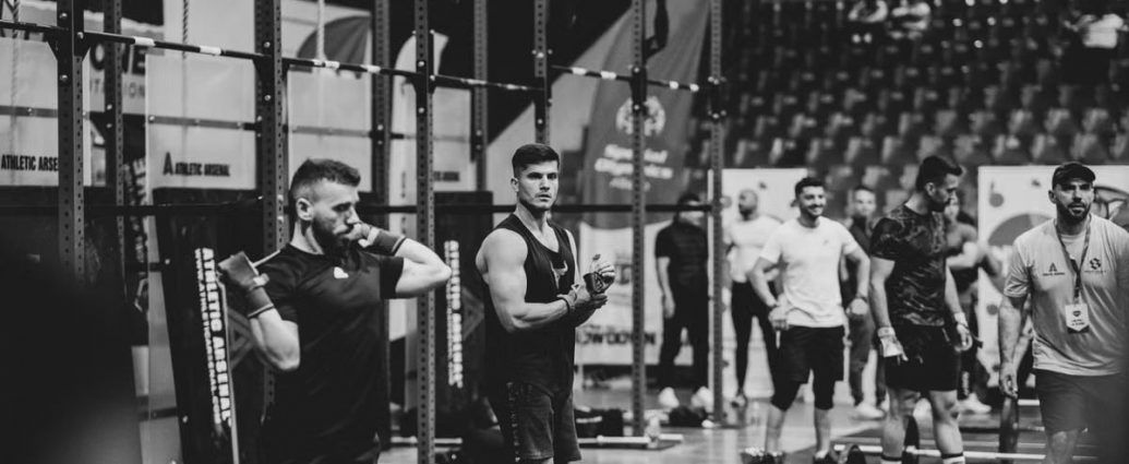 Black and white image of a fitness competition showcasing athletes and weights in a gym setting.